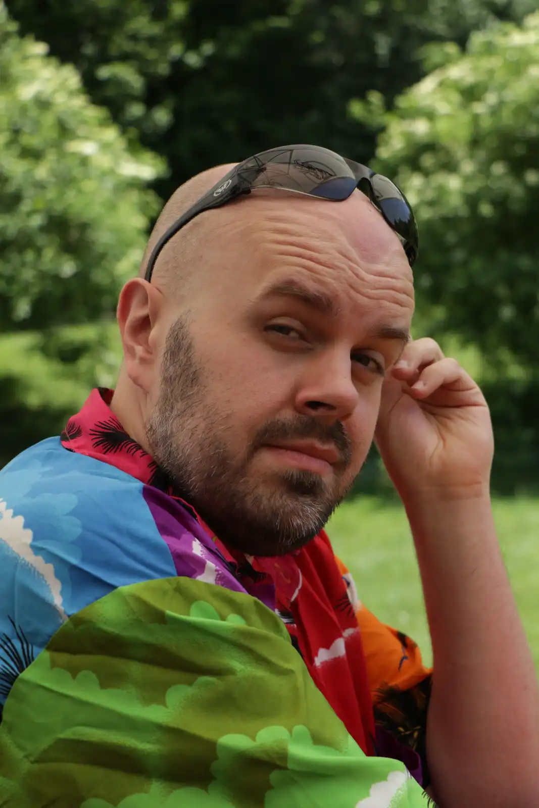 A bald man with a beard and sunglasses perched on his head looks directly at the camera with a weary expression, his hand…