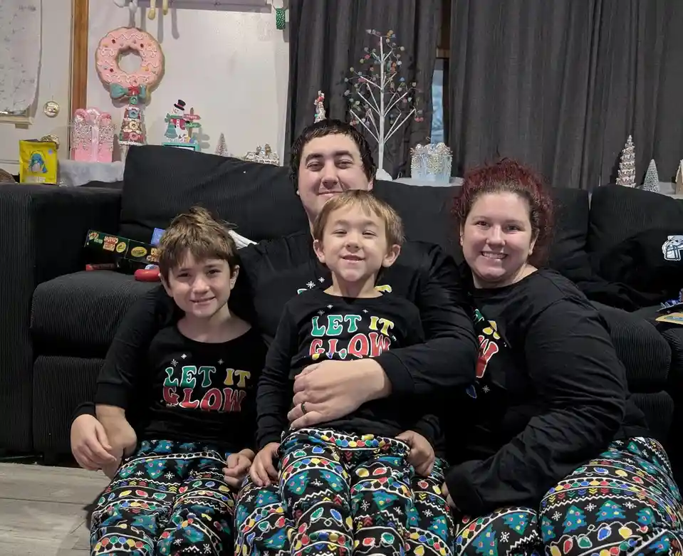 A family of four—two adults and two young boys—are sitting together on a black couch in a decorated living room. They are…