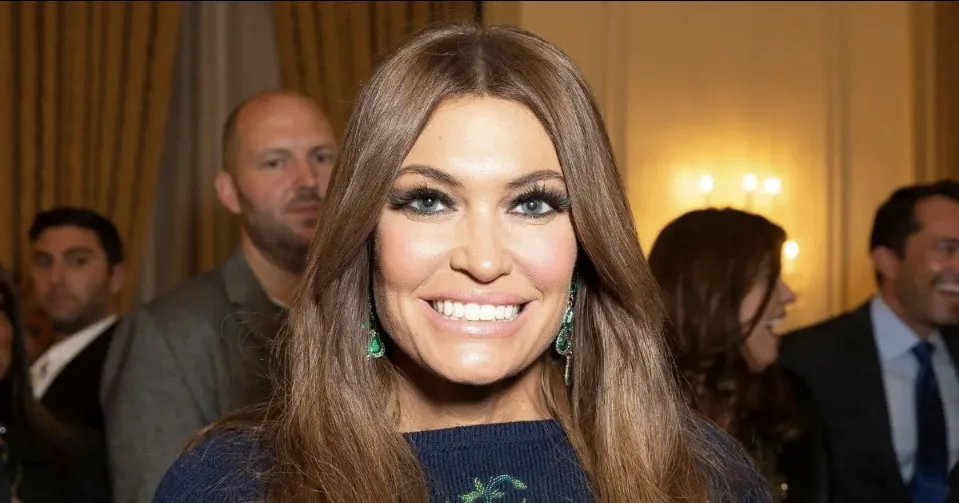 A woman with long brown hair and bright green earrings smiles widely at the camera while wearing a dark blue sweater. Behind…