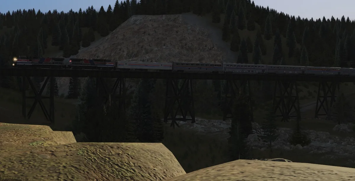 A long Amtrak-style passenger train crosses a wooden trestle bridge through a forested mountain valley at dusk. The image…