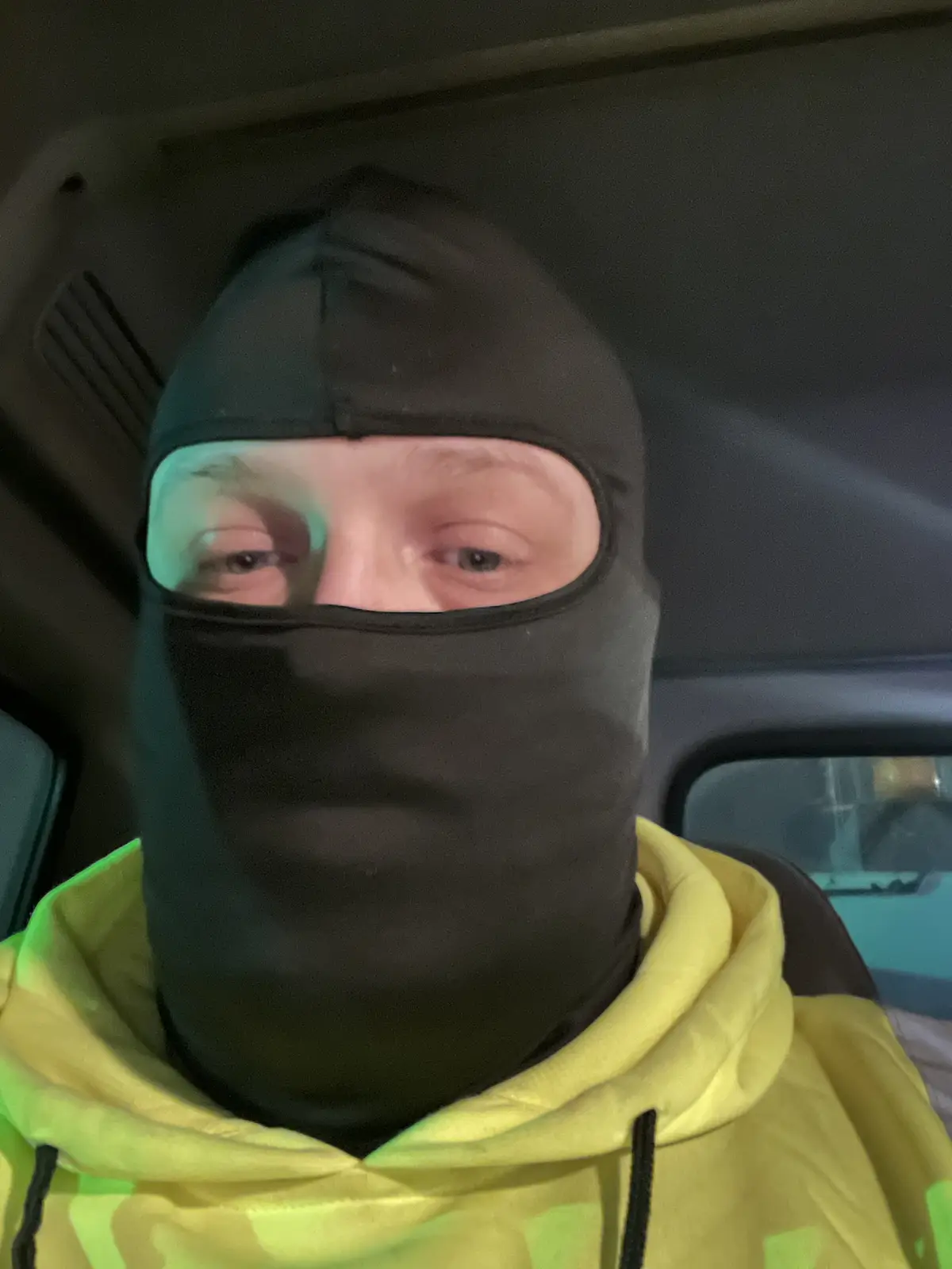 A person wearing a black balaclava covering their entire face except for their eyes, seated inside a car. They are wearing a…