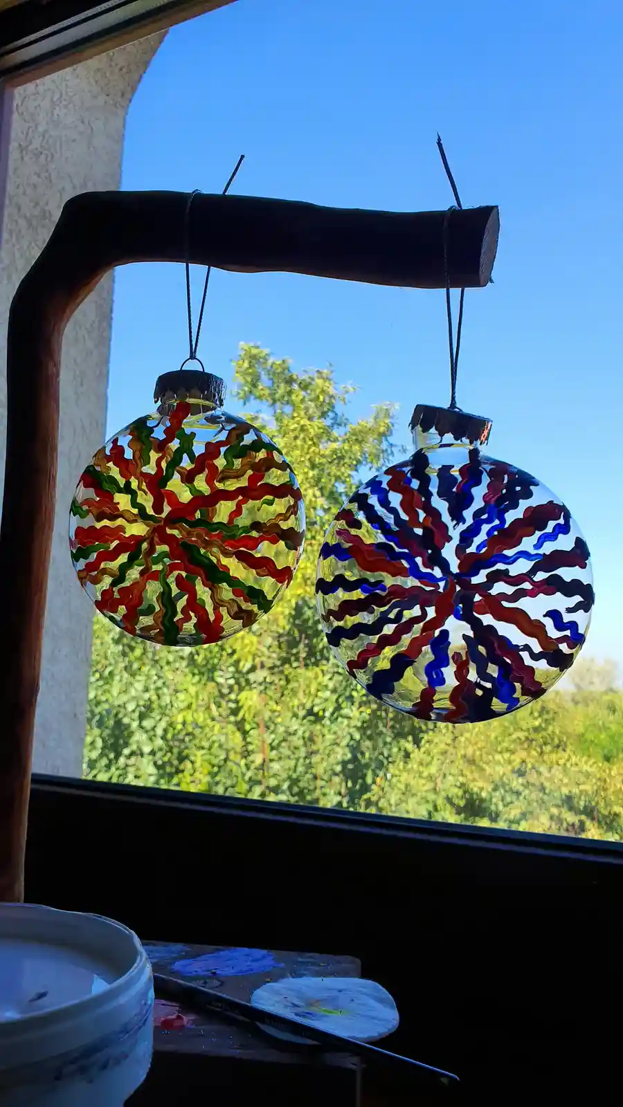 Two hand-painted glass ornaments hang from a wooden branch against a window, showing vibrant wavy patterns of red, green…