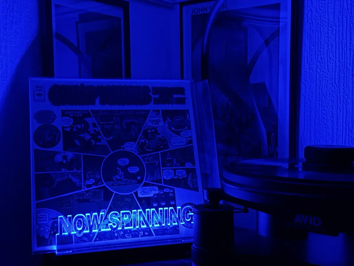 A vinyl record sleeve for 'Captain Beefheart & His Magic Band' is displayed in a dim blue-lit room next to a turntable. The…