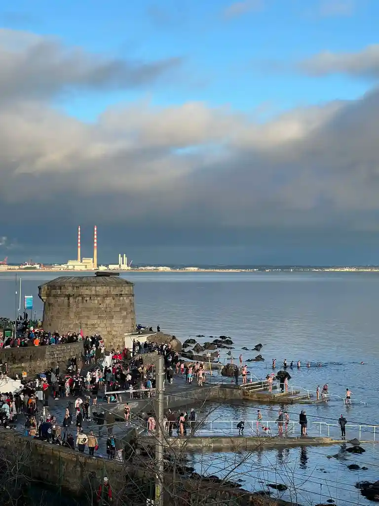 A wide shot shows a large crowd of people gathered at a seaside location with a stone tower and steps leading into the…