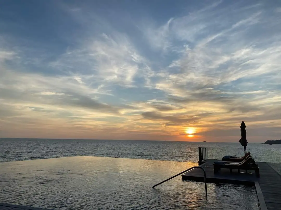A serene sunset over an infinity pool with lounge chairs and an umbrella silhouetted against the horizon. The overlaid text…