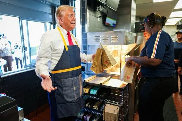 A man in a white shirt, red tie, and blue apron with a yellow stripe stands in a fast-food kitchen, gesturing with his left…
