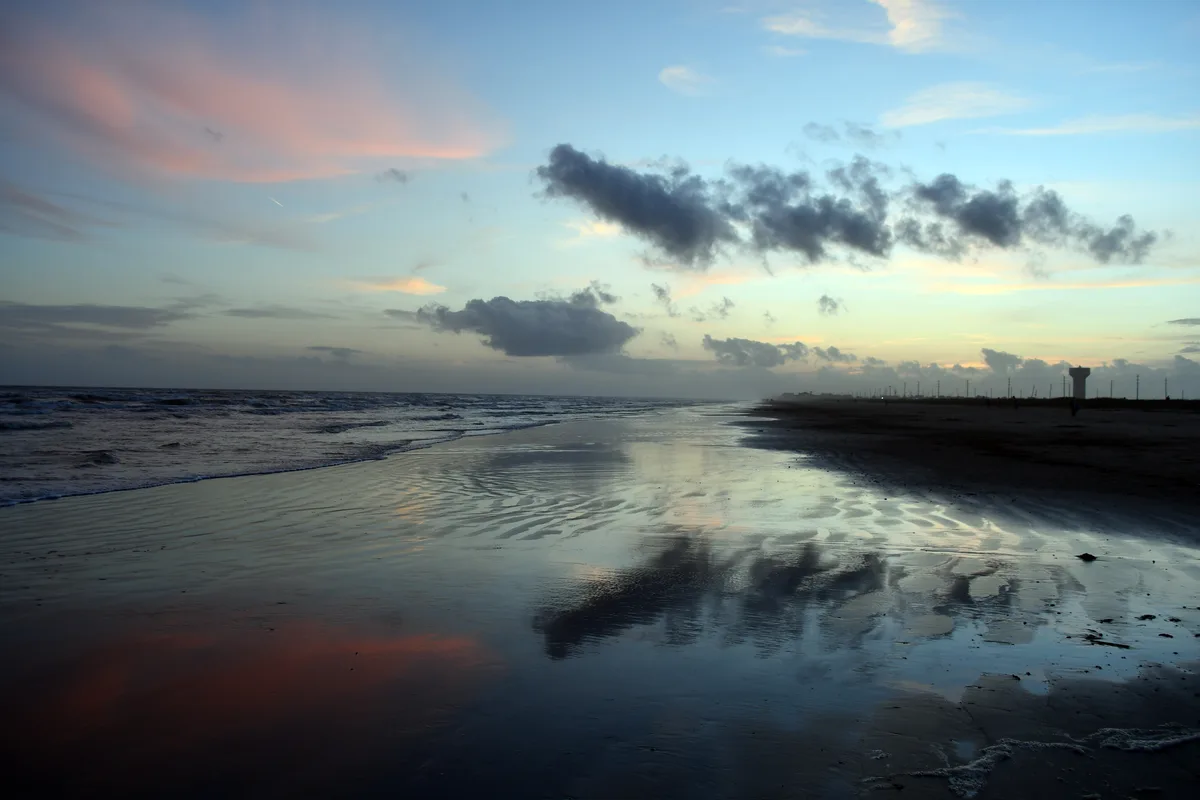A serene beach at sunset with pink and blue skies reflected in wet sand, distant clouds, and a lighthouse silhouette…