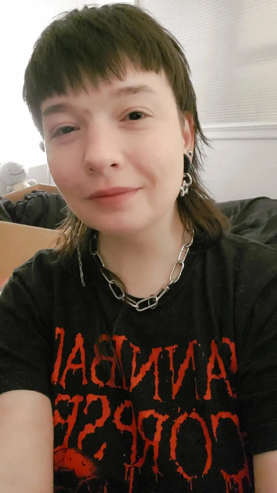 A person with short dark hair and a subtle smile wears a black t-shirt with the band name 'Cannibal Corpse' printed in…