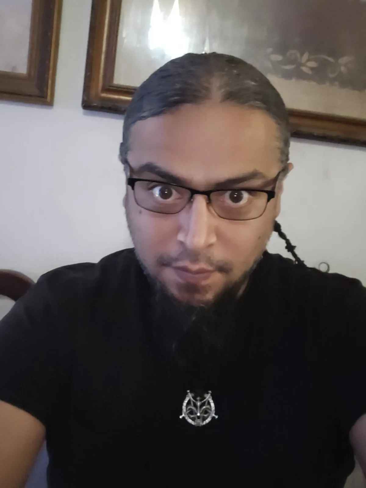 A man with glasses, a beard, and long hair pulled back in a braid stares intensely at the camera with wide eyes and a…