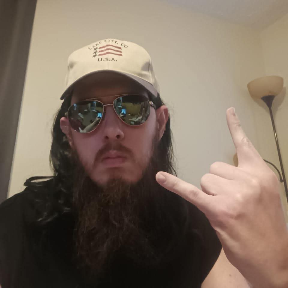 A man with long dark hair and a full beard wears a beige baseball cap with 'LAKE CITY, CO U.S.A.' and a stylized American…