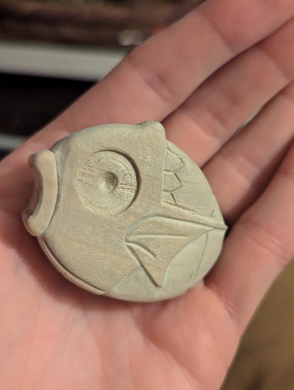 A hand holds a small, light-colored, carved wooden pendant shaped like an owl, featuring a circular eye, ear tufts, and…