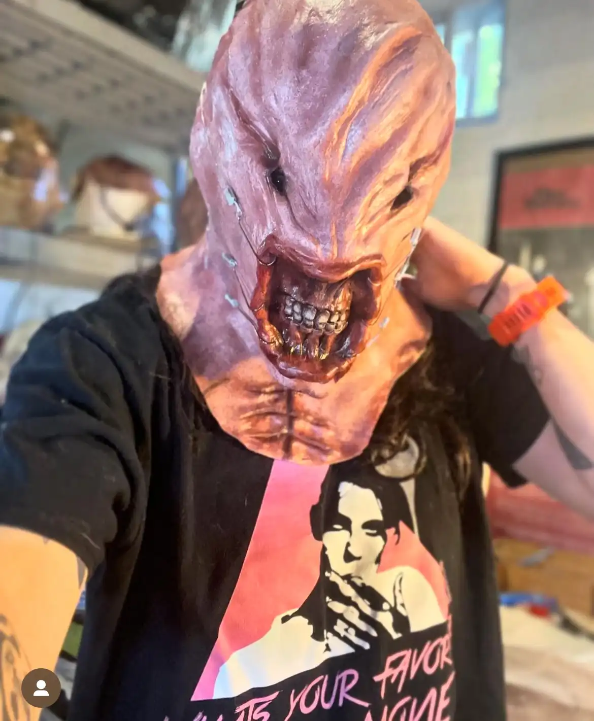 A person wearing a grotesque, detailed horror mask with a wide, toothy grin and exposed wires, while adjusting the mask with…