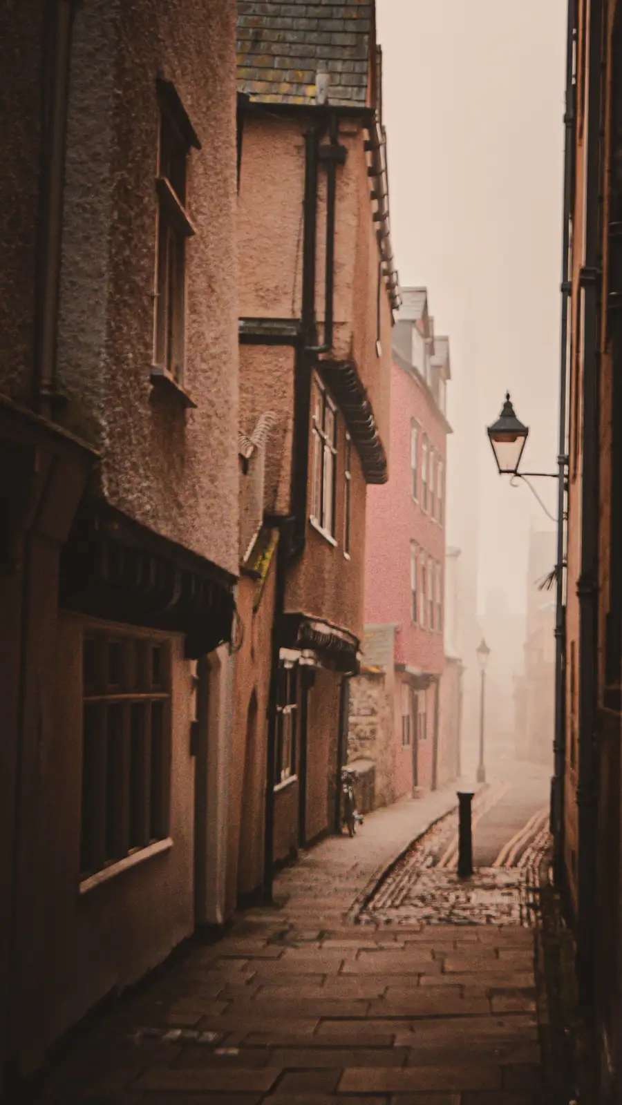 A narrow, foggy cobblestone alleyway between old brick buildings, with a vintage street lamp on the right and a bicycle…