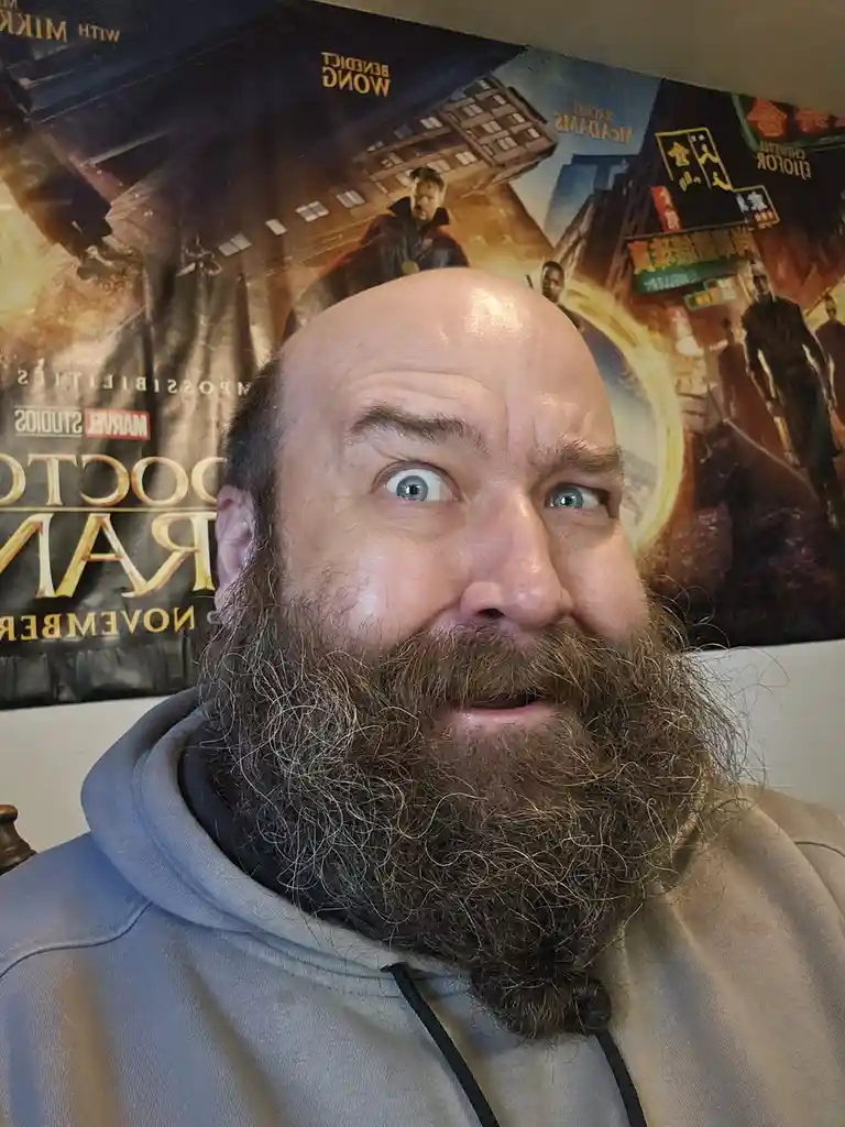 A man with a large, full beard and a bald head stares with wide, surprised eyes at the camera, standing in front of a…