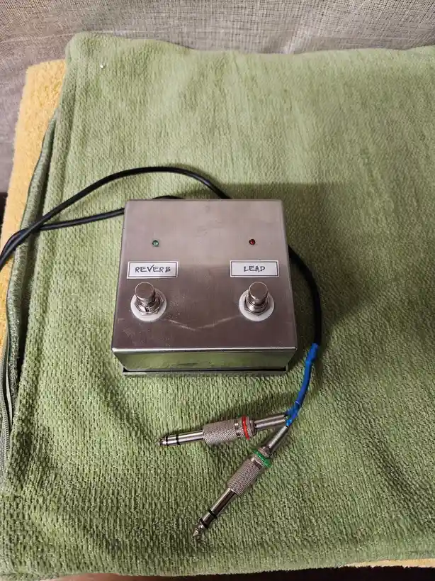 A custom-built metal guitar effects pedal with two footswitches labeled 'REVERB' and 'LEAD', sitting on a green towel. The…