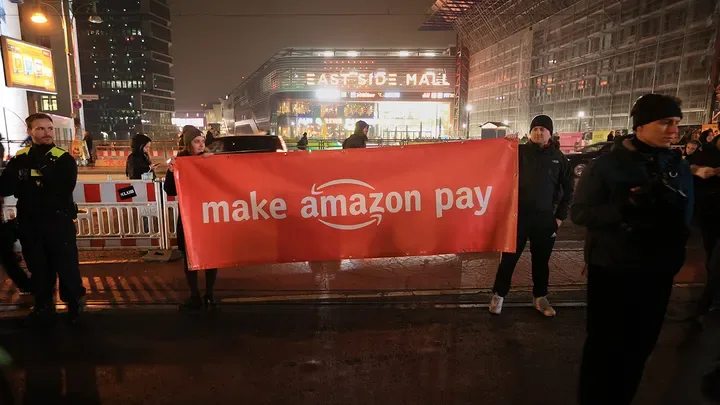 At night, protesters hold a large red banner with white text reading 'make amazon pay' in front of a brightly lit 'EAST SIDE…
