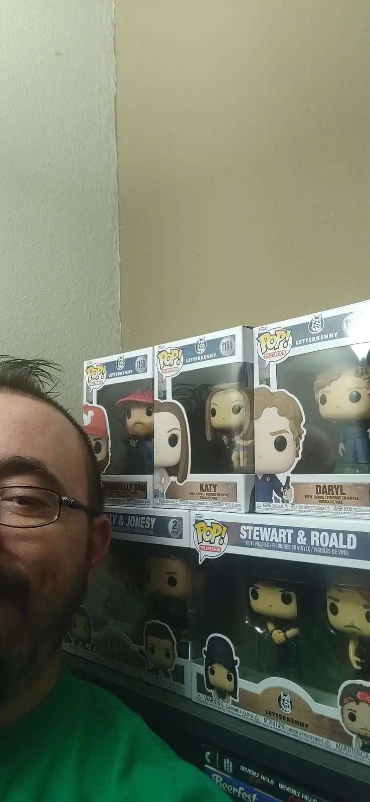 A man in a green shirt and glasses smiles at the camera while standing beside shelves displaying multiple Funko Pop! vinyl…