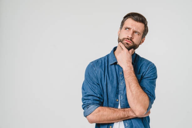 a man with a beard wearing a blue denim shirt stands against a plain white background, looking upward and to his right with…