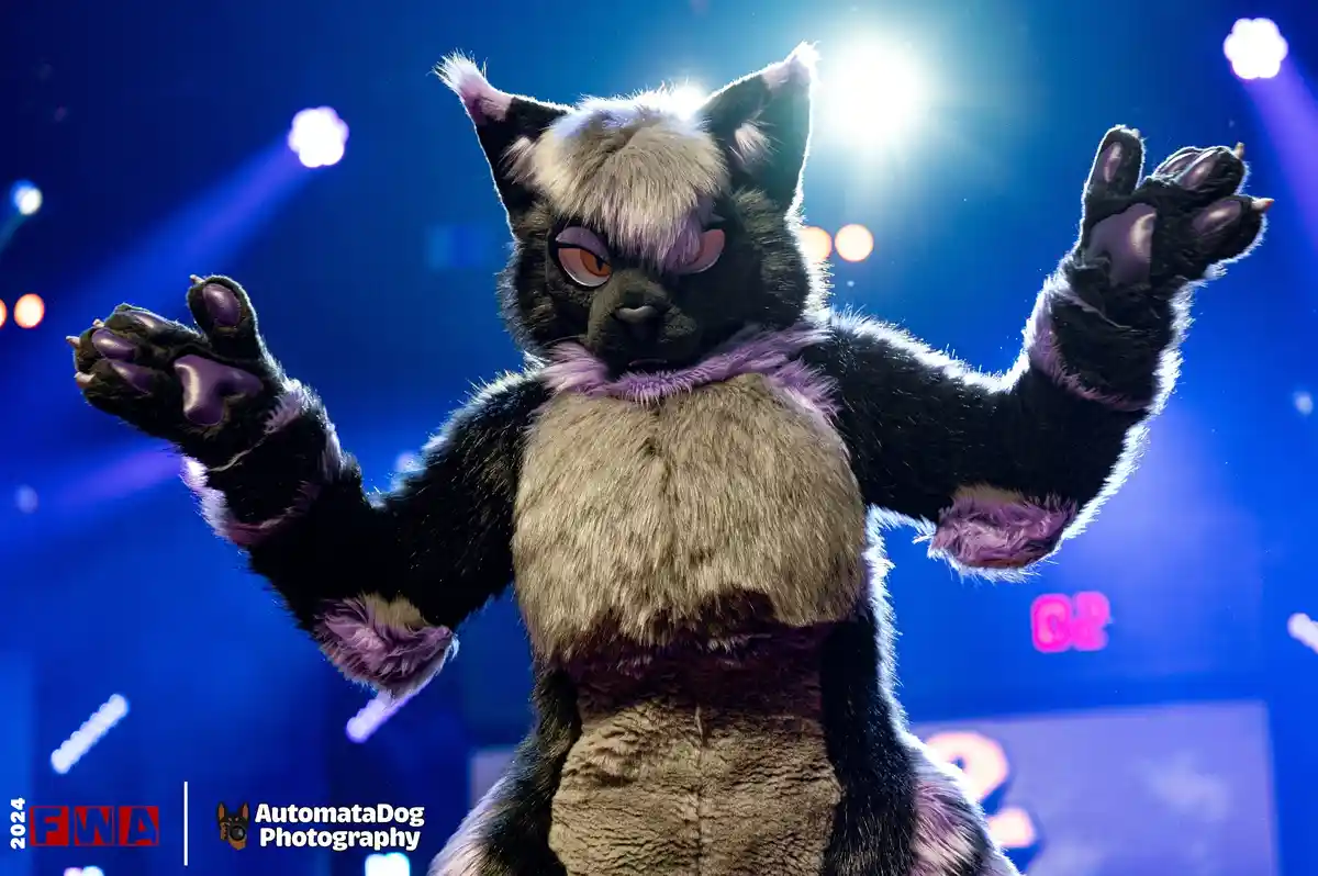 A person in a detailed black, grey, and purple fursuit resembling a cat stands on a stage under bright blue lights with arms…