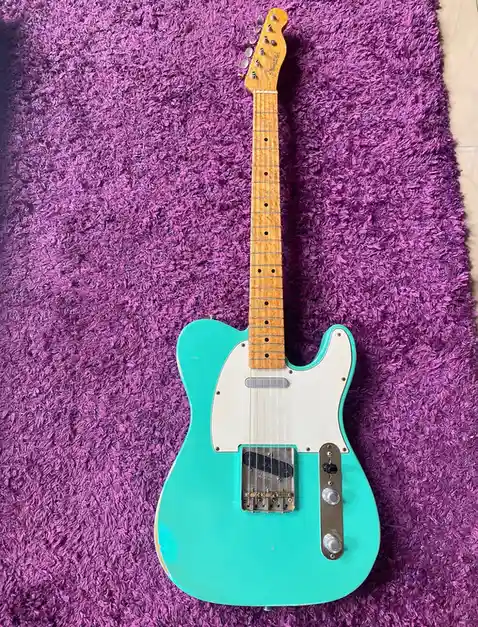 A turquoise Fender Telecaster electric guitar with a maple neck and white pickguard lies flat on a plush, shaggy purple rug…