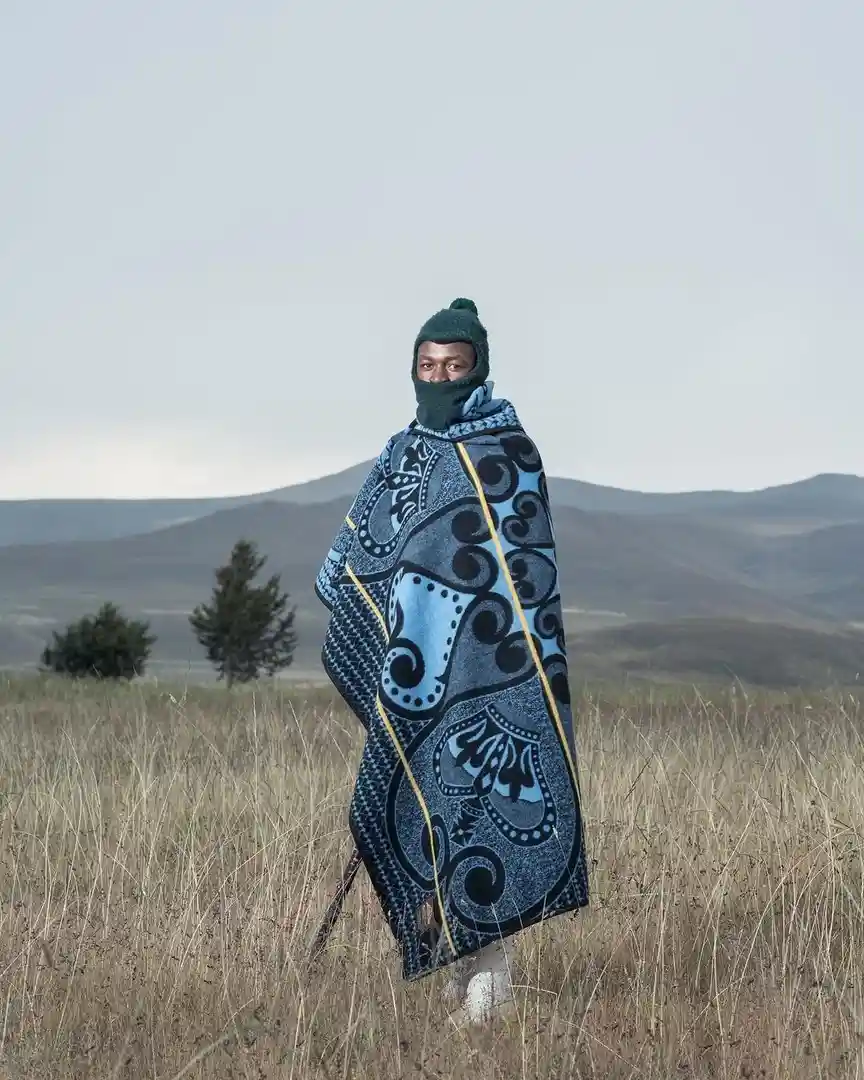 a person stands in a field of tall dry grass with rolling hills in the background, wearing a green balaclava and a large…