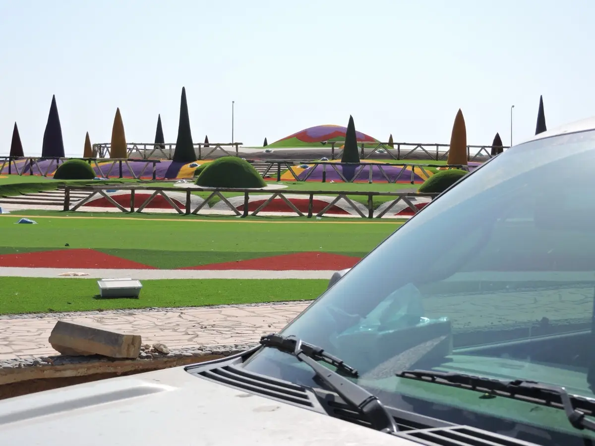 A photo taken from inside a car shows a brightly colored, artificial landscape with geometrically shaped hedges, painted…
