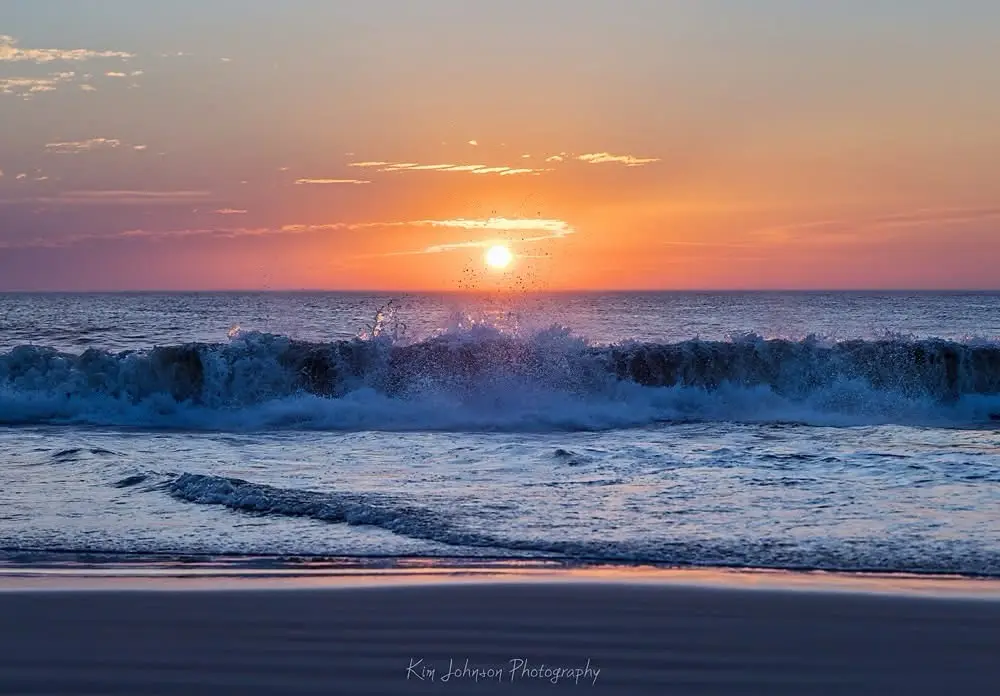 A serene sunset over the ocean with waves crashing in the foreground, the sun low on the horizon casting orange and pink…