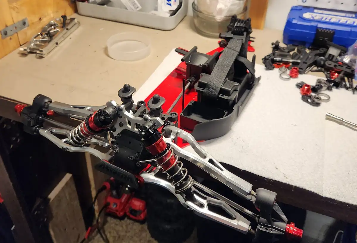 A close-up photo of a partially assembled red and silver RC car chassis on a workbench, surrounded by scattered black and…