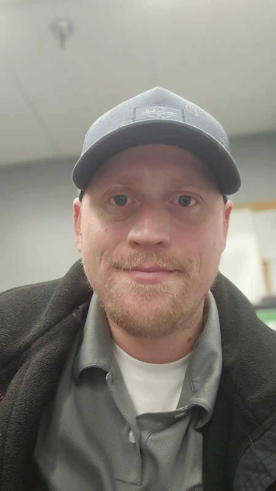 A man with a light beard and blue eyes wears a grey baseball cap and a grey polo shirt under a dark fleece jacket. He looks…