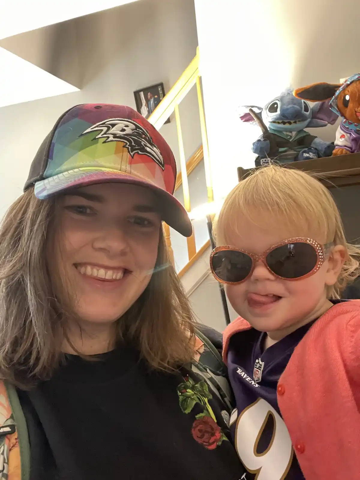 A woman wearing a rainbow-patterned Baltimore Ravens baseball cap smiles while holding a toddler who wears oversized…