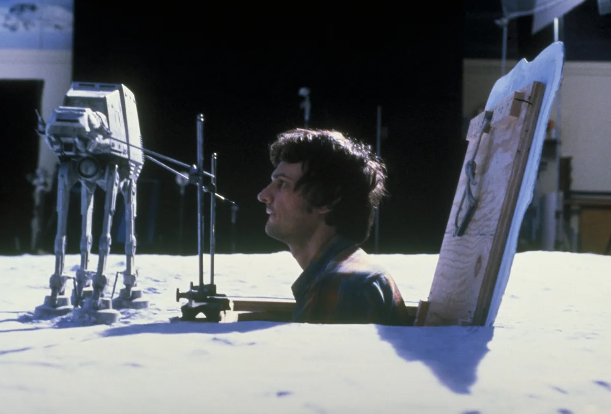 A man sits in a snowy, studio-like set, partially buried in white material, looking toward a model of an AT-AT walker. He is…