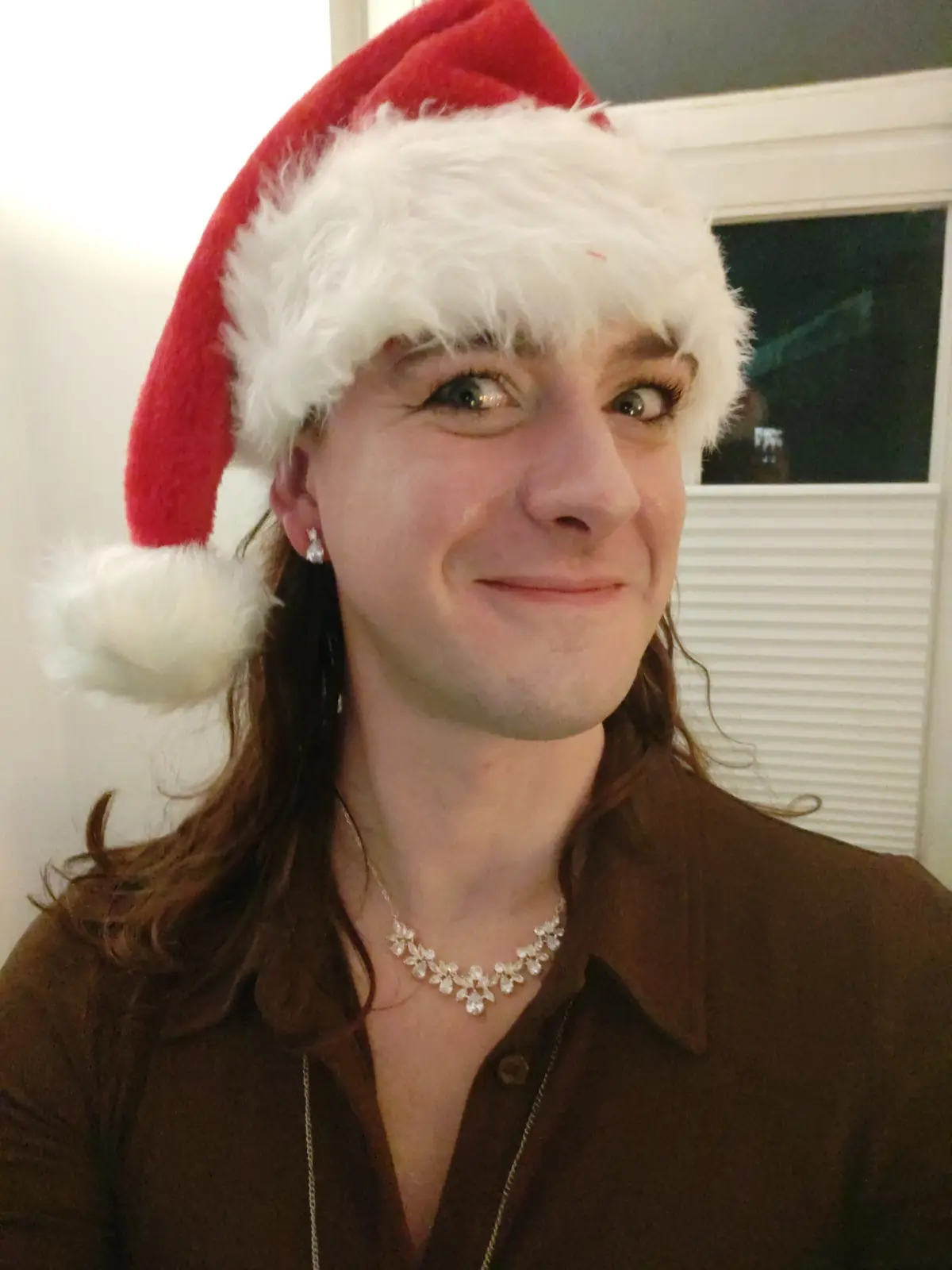 A person with long brown hair wears a red and white Santa hat, a brown collared shirt, and a floral crystal necklace…