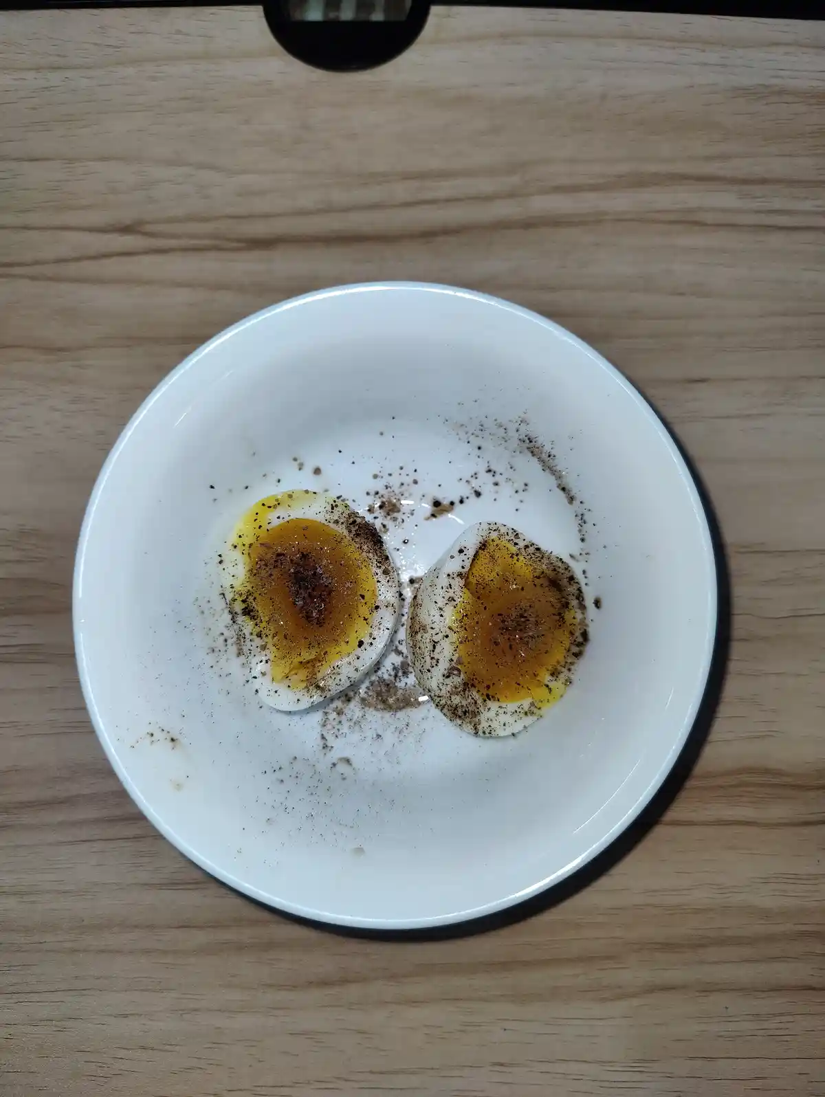 A top-down photo shows a white plate on a wooden surface holding two halves of a hard-boiled egg, seasoned with black…