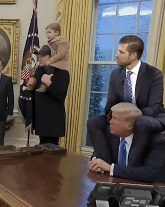 A still image from a White House event shows a man in a dark suit holding a small child on his shoulders, while another man…