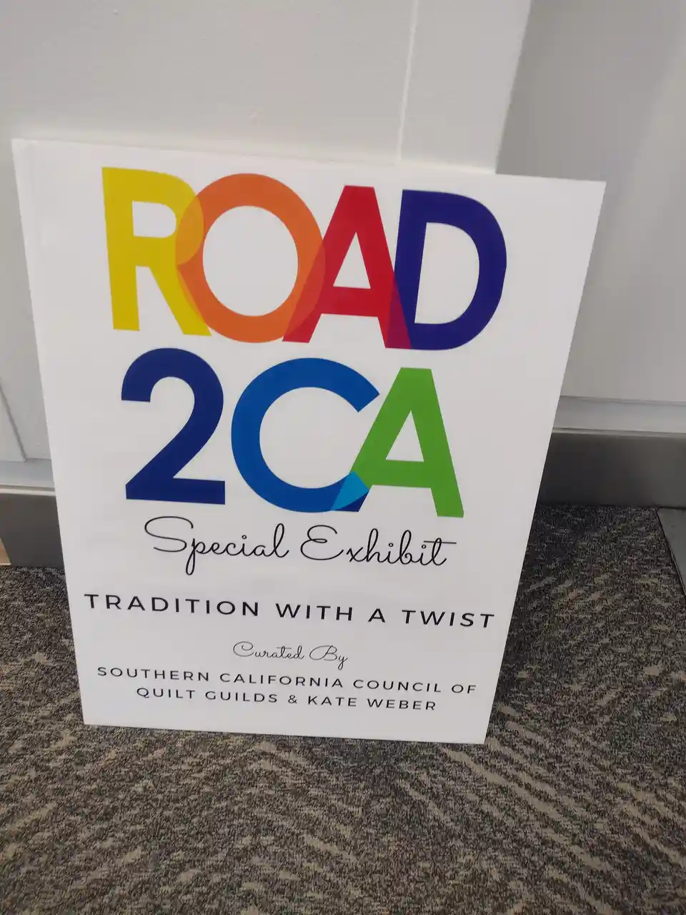 A white sign stands on a patterned carpet against a wall, advertising a 'ROAD 2CA Special Exhibit' with the tagline…