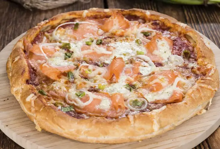a close-up photo of a round, thick-crust pizza on a wooden board, topped with melted cheese, slices of pink salmon, chopped…