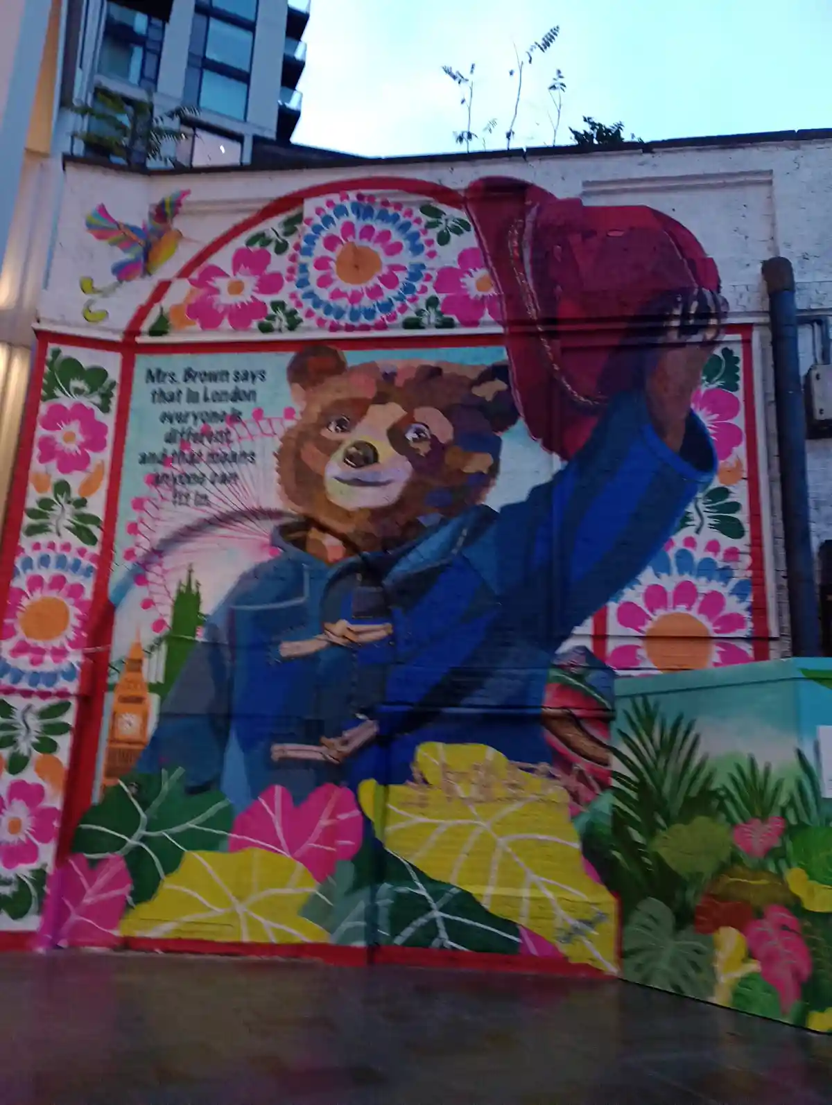 A colorful mural depicts Paddington Bear wearing his signature blue coat and red hat, standing amidst vibrant floral…