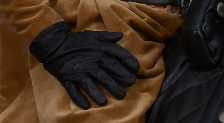 A close-up shot shows a black leather glove resting on a brown velvet fabric, with part of a black leather garment visible…
