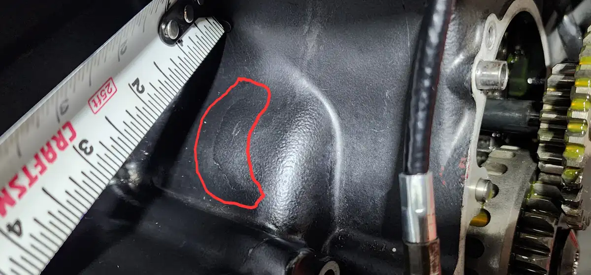 A close-up photo of a black engine block with a red outline highlighting a shallow dent. A white measuring tape labeled…