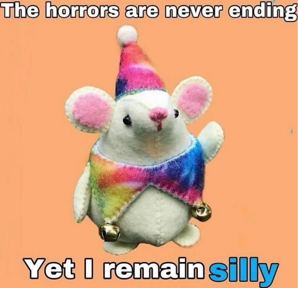 A plush white mouse toy wearing a rainbow tie-dye party hat and matching scarf with bells, set against a solid peach…