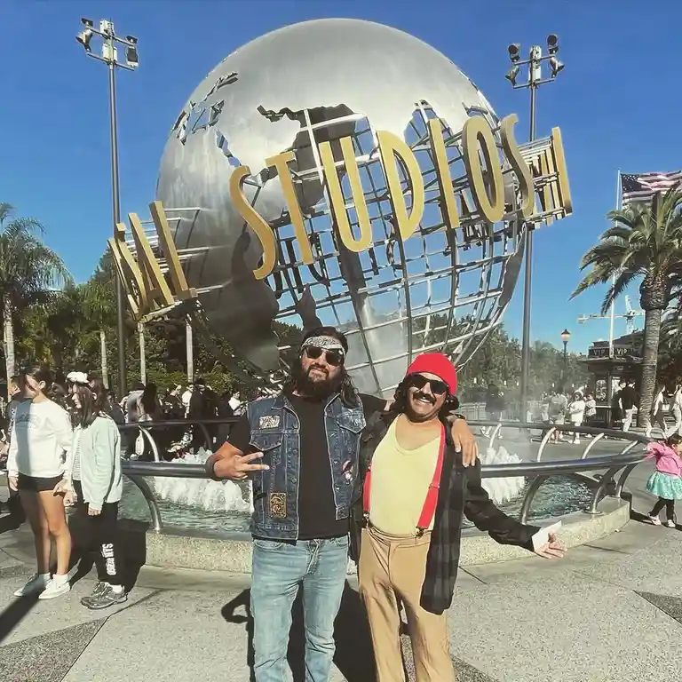 Two men posing in front of the Universal Studios globe landmark, one dressed in a denim vest and bandana, the other in a red…