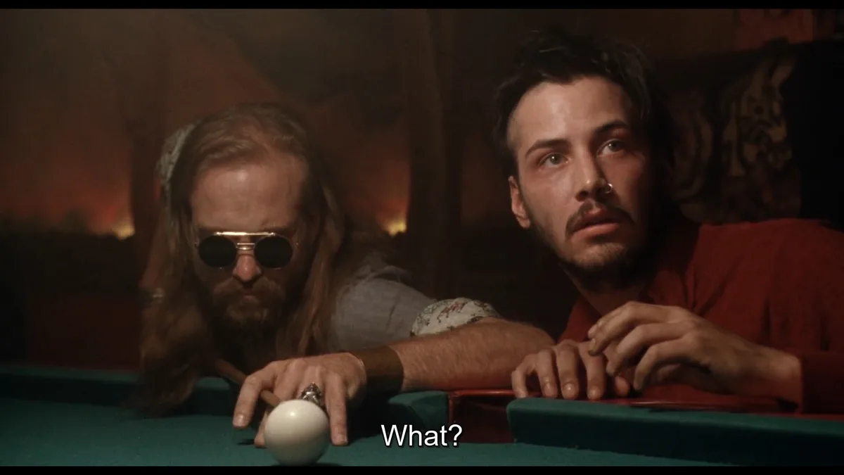 Two men are leaning over a green pool table in a dimly lit room. The man on the left, with long hair and round sunglasses…
