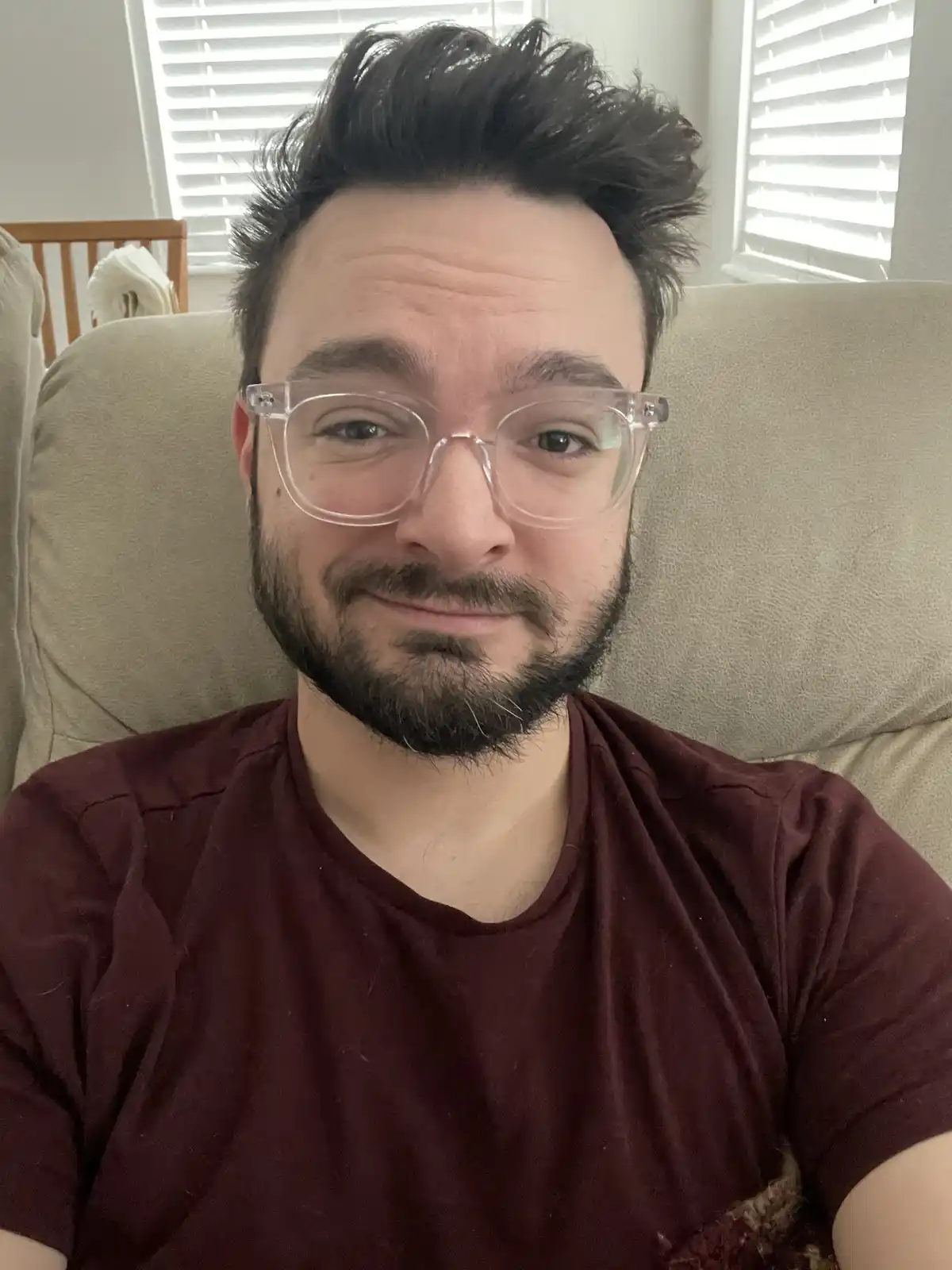 A man with dark hair, a beard, and clear-framed glasses sits on a beige couch, looking directly at the camera with a subtle…