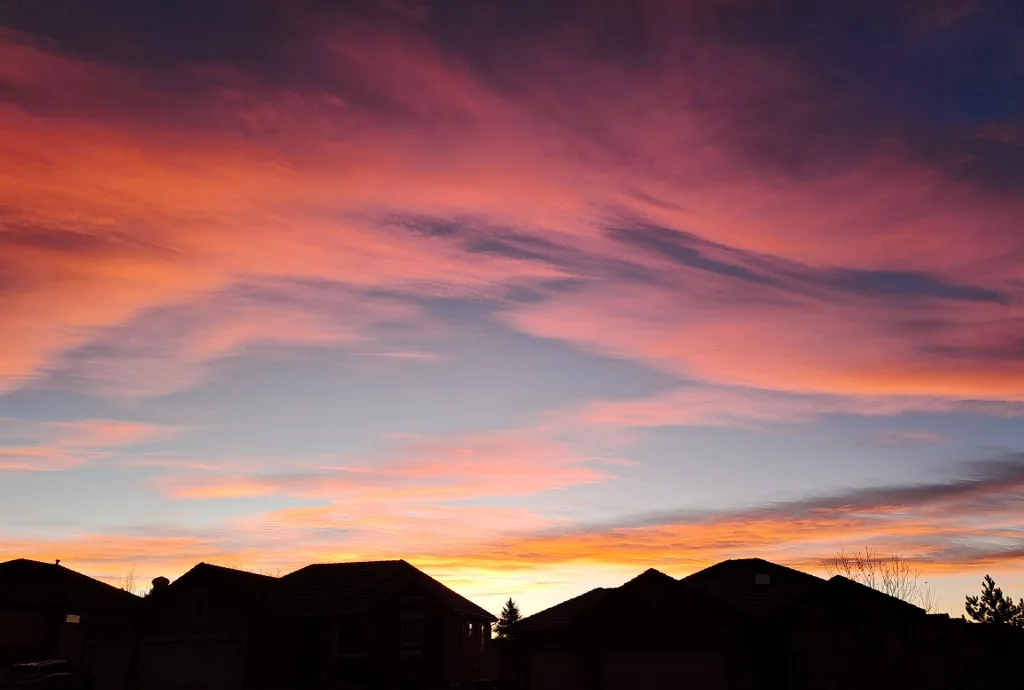 A vibrant sunset with fiery orange, pink, and purple clouds above the silhouettes of suburban houses. The overlaid text…