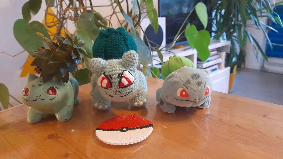 Three Bulbasaur-themed items sit on a wooden table: a ceramic planter with a succulent, a crocheted Bulbasaur wearing a…