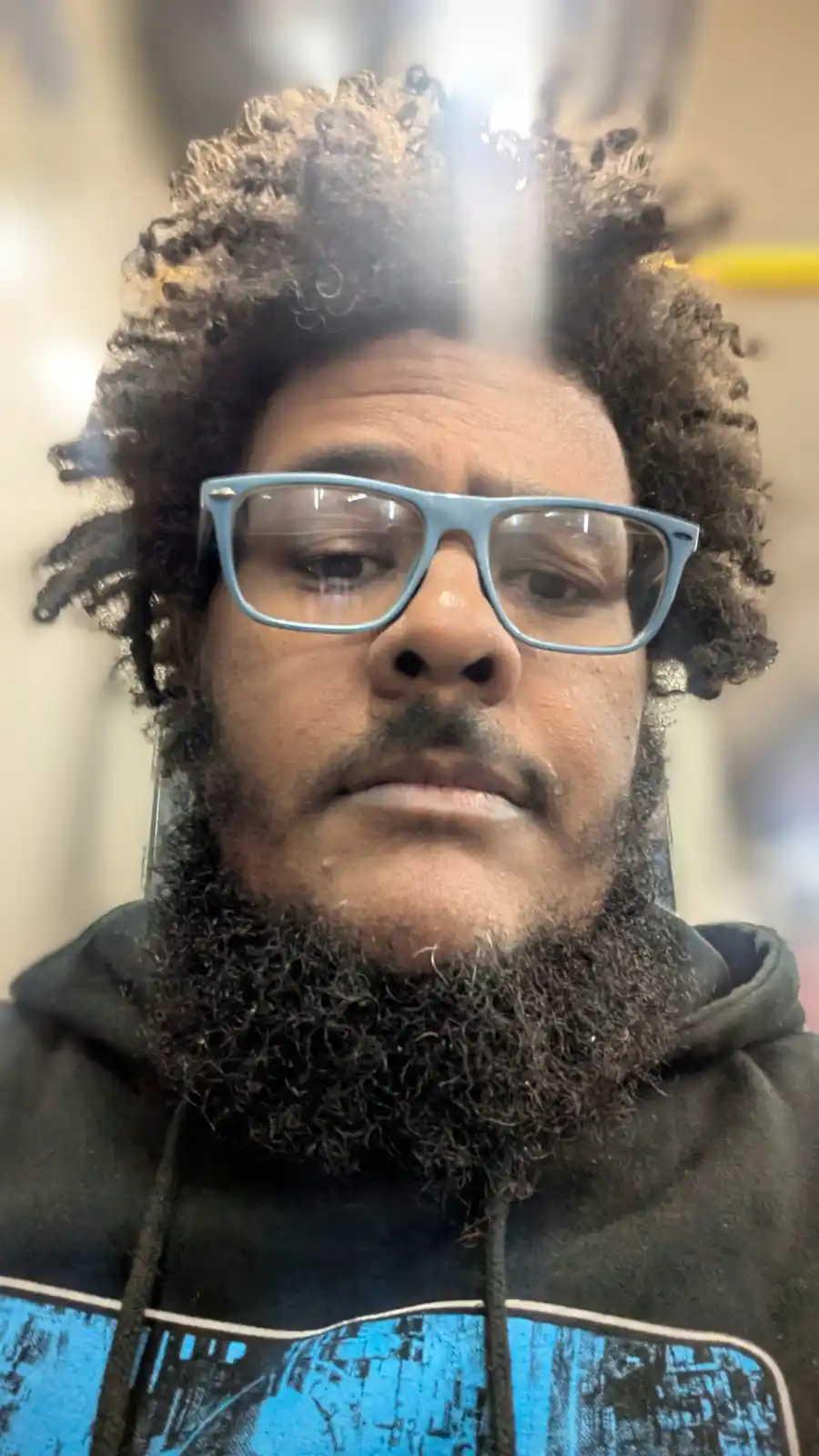 A close-up photo of a man with curly hair, glasses, and a beard, looking directly at the camera with a neutral expression. A…
