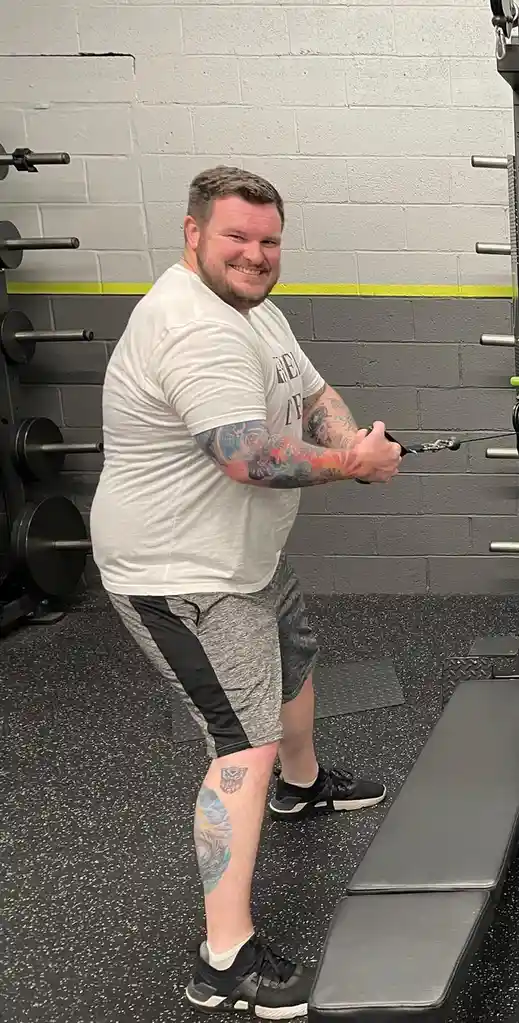 A smiling, tattooed man in a gym is using a cable machine, looking at the camera with a confident grin. He wears a white…