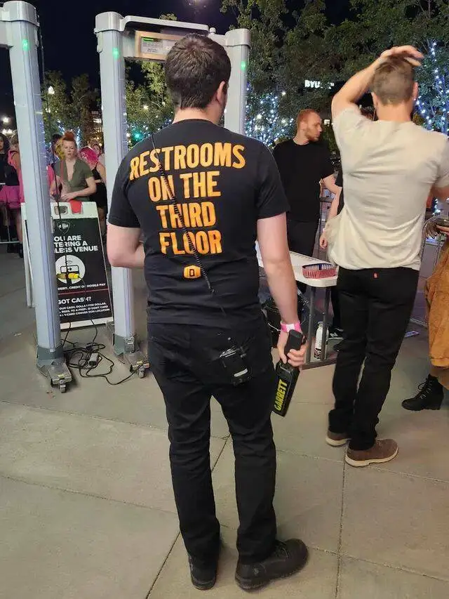A security guard at an event, seen from behind, wears a black t-shirt with large orange text reading 'RESTROOMS ON THE THIRD…