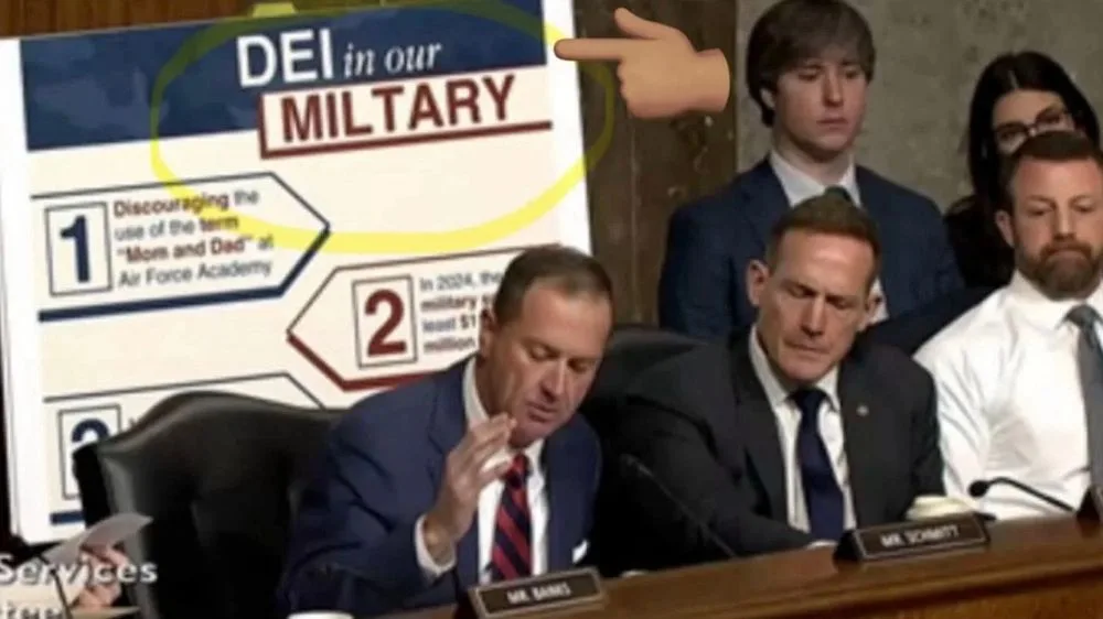 A man in a suit sits at a hearing table, looking down and rubbing his face, while a cartoon hand points to a poster behind…
