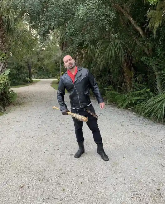 A man in a black leather jacket, red shirt, and dark pants stands on a gravel path surrounded by lush greenery, holding a…